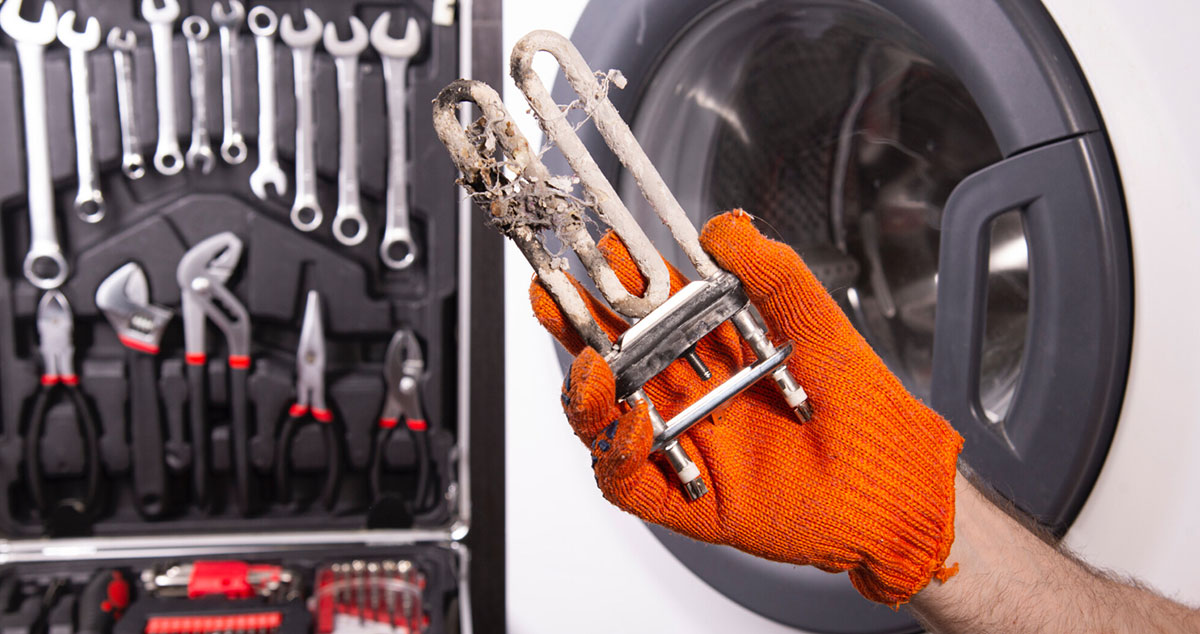 Eine Hand mit Handschuh hält ein verkalktes Ersatzteil vor einer Waschmaschine und einem Werkzeugkoffer.